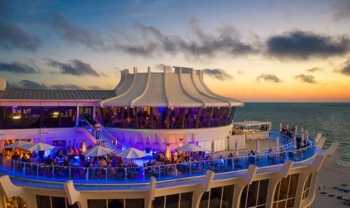 A vibrant rooftop restaurant with outdoor seating, overlooking the ocean at sunset, illuminated by colorful lights.