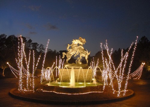 A illuminated fountain with a statue of horses, surrounded by twinkling lights and trees at dusk.