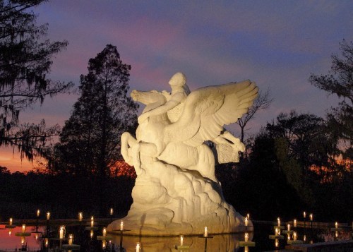 A white statue of a winged figure atop a horse, surrounded by candles, against a colorful sunset sky.