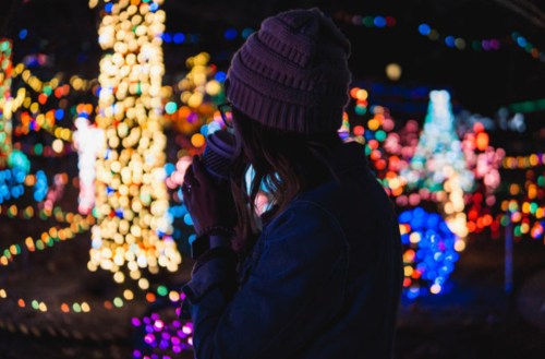 A person in a beanie holds a cup, surrounded by colorful holiday lights in a festive outdoor setting.