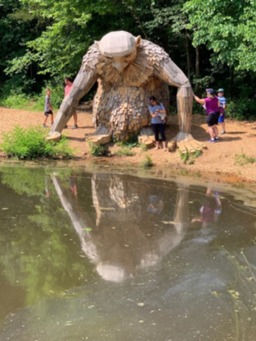 A large wooden sculpture of a creature by a pond, with people interacting nearby and its reflection in the water.