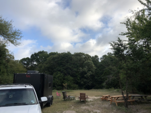 A campsite with a white vehicle, a black trailer, picnic tables, and trees under a partly cloudy sky.