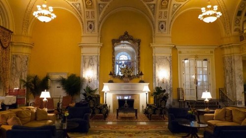 Elegant hotel lobby with yellow walls, marble columns, a fireplace, and plush seating under ornate chandeliers.
