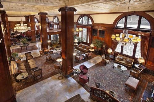 Elegant hotel lobby featuring wooden pillars, plush seating, and large windows, adorned with chandeliers and decorative rugs.