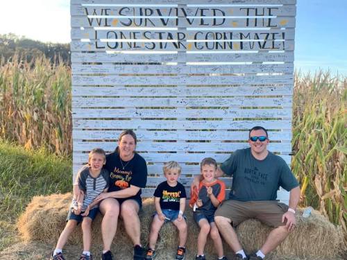 A family with three young boys sits on hay bales in front of a sign that reads "We Survived the Lonestar Corn Maze!"