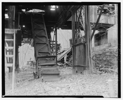 An abandoned industrial site featuring a metal staircase and dilapidated structures surrounded by overgrown vegetation.