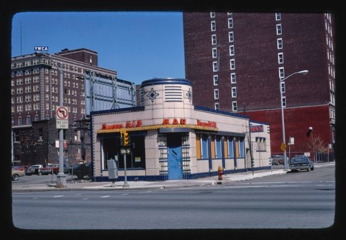 A vintage diner with a blue and cream facade, located at a street corner, surrounded by buildings and parked cars.