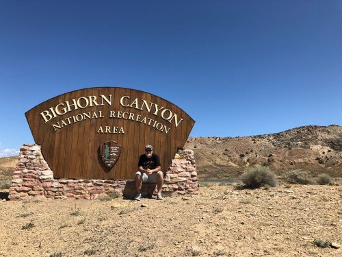 A person sits next to the Bighorn Canyon National Recreation Area sign, with a desert landscape and blue sky in the background.