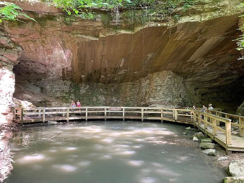 A wooden walkway curves around a serene pond inside a rocky cave, surrounded by lush greenery.