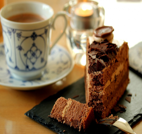 A slice of chocolate cake with chocolate shavings, next to a cup of tea on a wooden table.