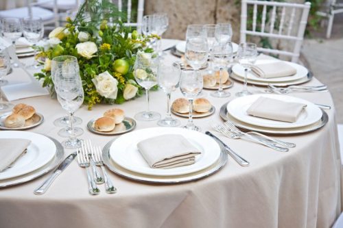 Elegant dining table set with white plates, glassware, napkins, and a floral centerpiece. Rolls are placed on small plates.