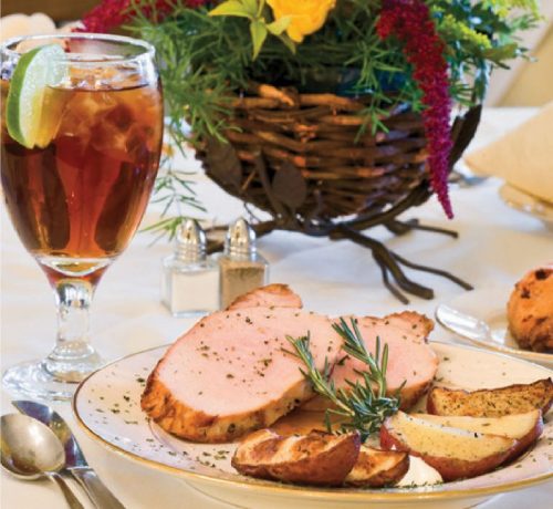 A plate of sliced pork with roasted potatoes, garnished with rosemary, alongside a glass of iced tea and a floral centerpiece.