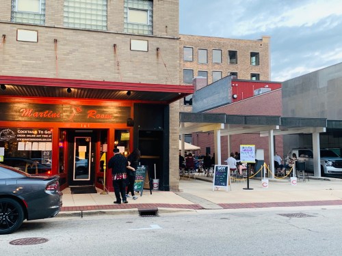 Exterior view of a bar with outdoor seating, people mingling, and a car parked nearby on a city street.