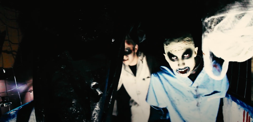 Two eerie figures with ghostly makeup and dark clothing emerge from a shadowy background, creating a chilling atmosphere.
