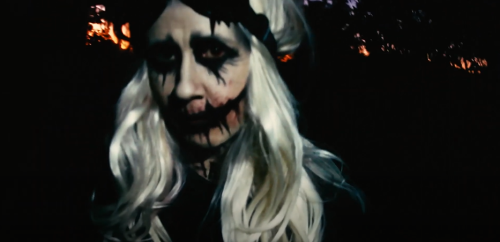 A person with pale skin and long white hair, wearing dark makeup and a haunting expression, stands against a dark background.