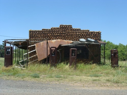 Abandoned gas station with rusted pumps, a crumbling structure, and overgrown grass under a clear blue sky.