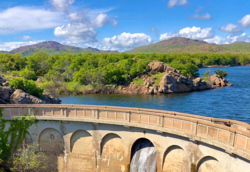 A scenic view of a dam with water flowing, surrounded by lush greenery and mountains under a blue sky with clouds.