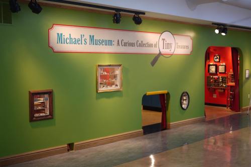 Sign for "Michael's Museum: A Curious Collection of Tiny Treasures" on a green wall, with display cases and a doorway.