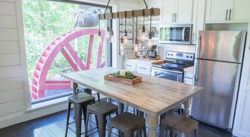 Bright kitchen with a wooden table, metal stools, and a large window showcasing a red waterwheel outside.