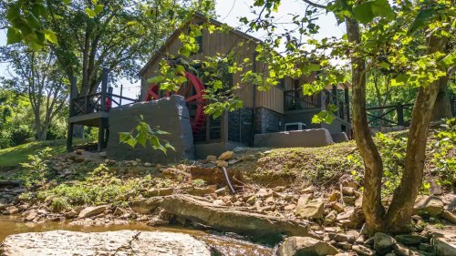 A rustic building with a large water wheel, surrounded by trees and a rocky stream in a sunny outdoor setting.