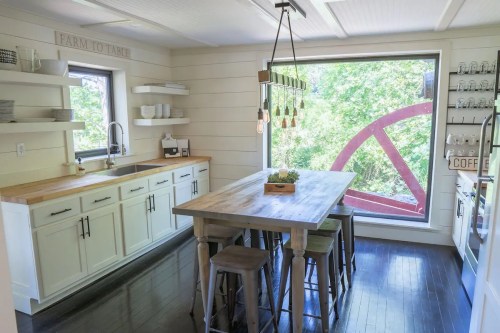 Bright kitchen with white cabinets, wooden table, and large window showcasing a scenic view with a red wheel outside.