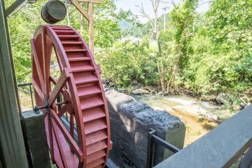 A red water wheel beside a flowing creek, surrounded by lush green trees and a sunny landscape.