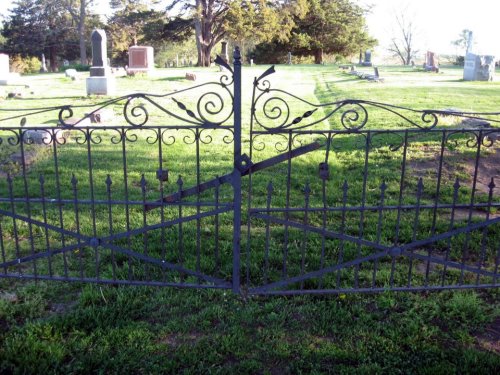 Wrought iron gate in a cemetery, surrounded by green grass and gravestones in the background.