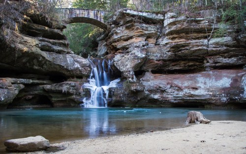 A serene waterfall cascades into a clear pool, surrounded by rocky cliffs and a stone bridge above.