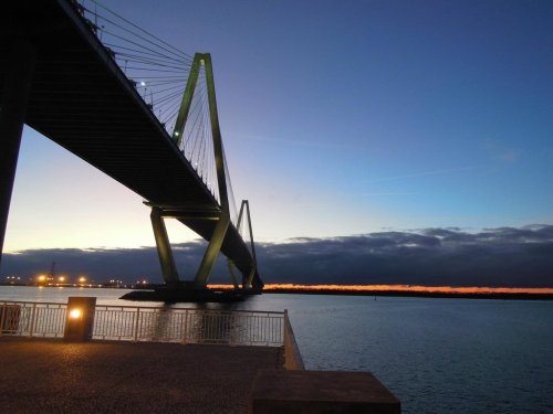 A bridge spans over water at dusk, with a colorful horizon and clouds in the background, reflecting lights on the surface.