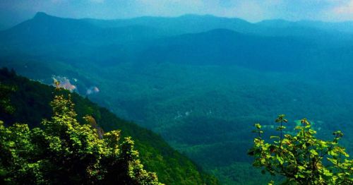 A panoramic view of lush green mountains under a hazy blue sky, showcasing layers of hills and valleys.