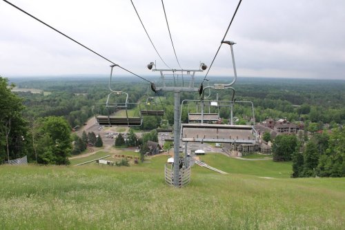 A ski lift ascends a green hillside, overlooking a resort and forested landscape under a cloudy sky.