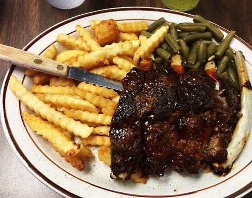 A plate of crinkle-cut fries, green beans, and a large piece of glazed meat with a knife resting on the side.