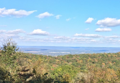 A scenic view of rolling hills and valleys under a blue sky with fluffy clouds, showcasing autumn foliage.