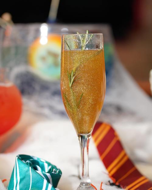 A sparkling cocktail in a tall glass, garnished with a sprig of rosemary, set against a festive background.