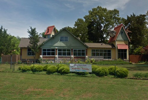 A colorful, two-story house with a green exterior and red accents, surrounded by a lawn and a sign for visiting hours.