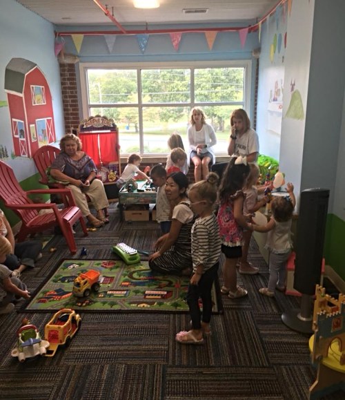 A colorful playroom filled with children playing with toys, while adults watch and interact in a bright, cheerful environment.