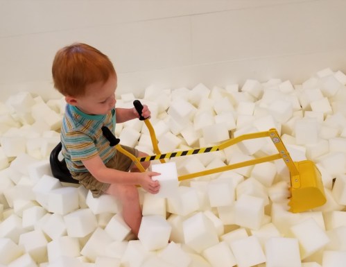 A young child sits on a pile of white foam blocks, using a yellow toy digger to scoop up a block.