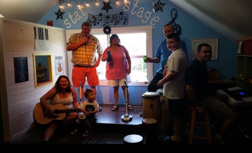 A family gathers on a small stage, with instruments, enjoying music and drinks in a colorful room.
