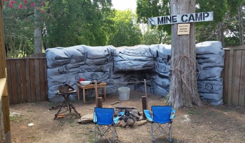 A rustic mine camp scene with a rock wall, campfire setup, and two blue chairs under a tree.