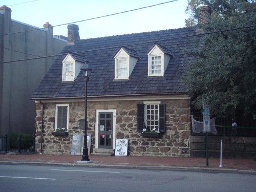 Historic stone building with a sloped roof, white windows, and a sign for a museum gift shop. Street view.