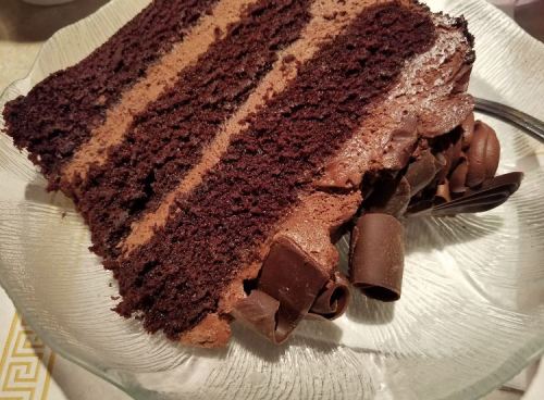 A slice of rich chocolate cake with layers of chocolate frosting and chocolate curls on a glass plate.