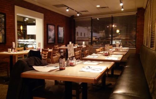 A dimly lit restaurant interior with empty tables, chairs, and condiments on the tables. Large windows with blinds.