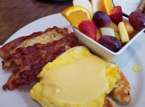 A plate with crispy bacon, a cheesy omelet, and a bowl of fresh fruit including strawberries, grapes, and orange slices.