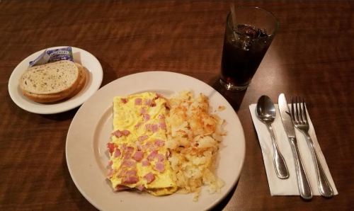 A plate with a ham and cheese omelet, hash browns, a glass of cola, and a side of bread on a table.