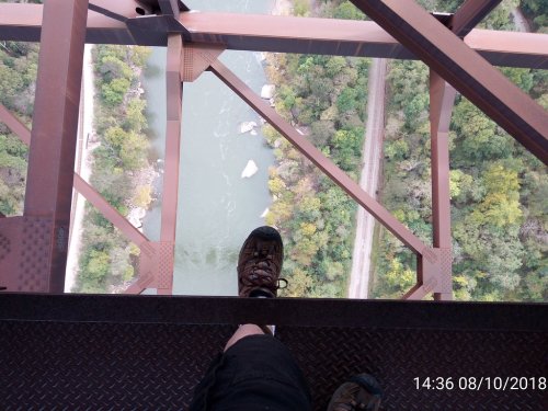 A person's foot on a metal beam above a river, with trees and a path visible below.