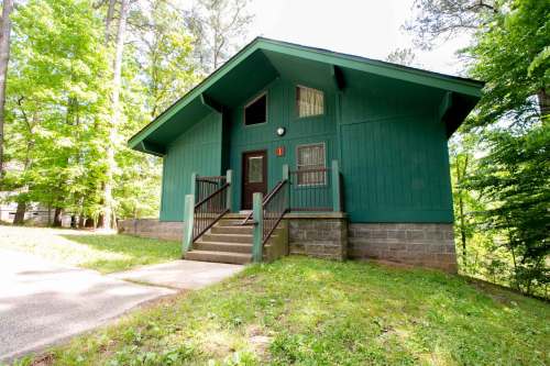A green, modern cabin with a sloped roof, surrounded by trees and grass, featuring a front staircase and porch.