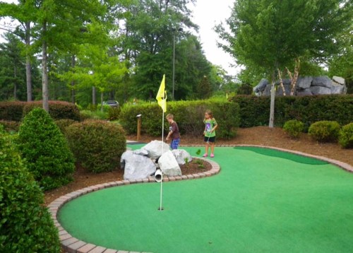 Two children play mini-golf on a green course surrounded by bushes and rocks, with a yellow flag in the background.