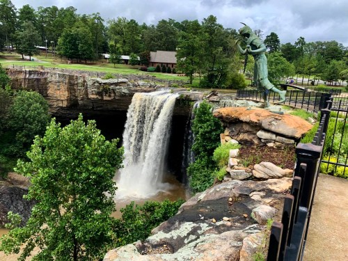 A statue of a man stands near a waterfall, surrounded by lush greenery and a scenic park setting.