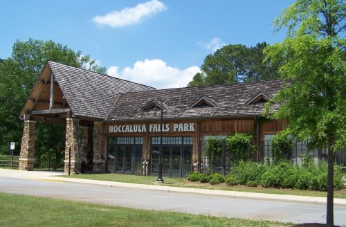 Entrance to Noccalula Falls Park featuring a wooden structure and stone pillars, surrounded by greenery and blue skies.