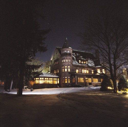 A grand, illuminated mansion surrounded by snow, set against a dark night sky with trees nearby.